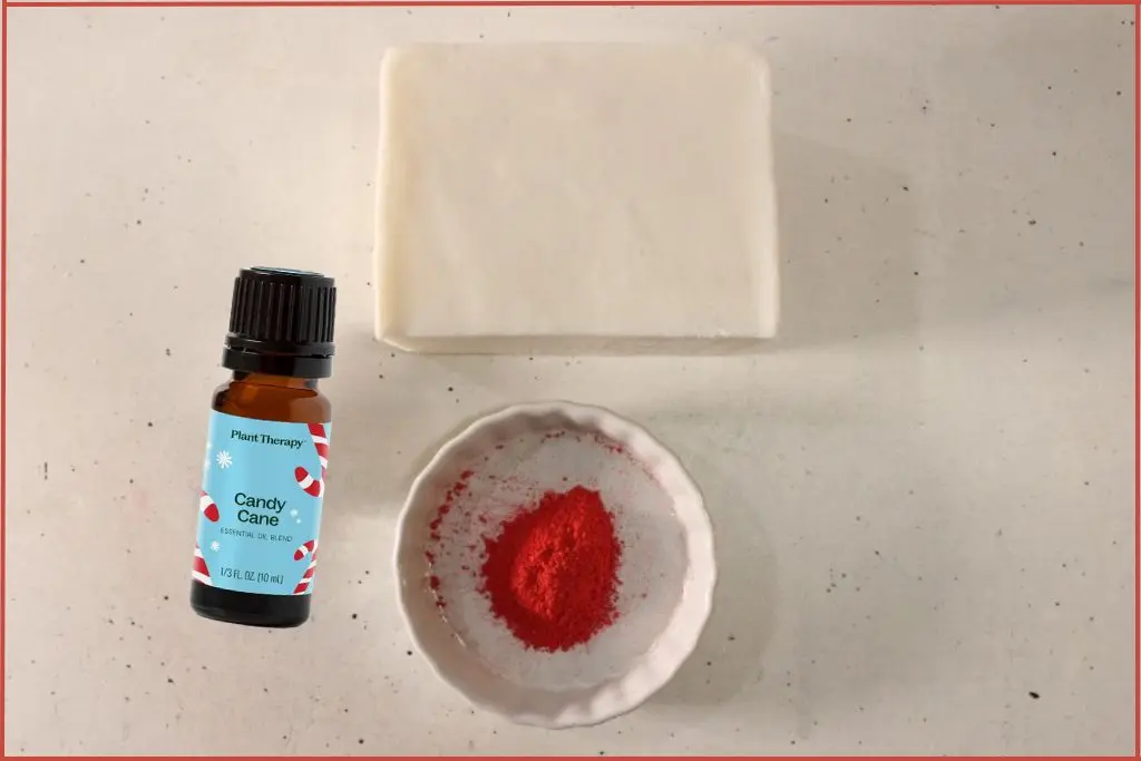 A block of goat's milk soap base, bowl with red mica powder, and bottle of candy cane essential oil blend - Ingredients required to make candy cane melt and pour soap