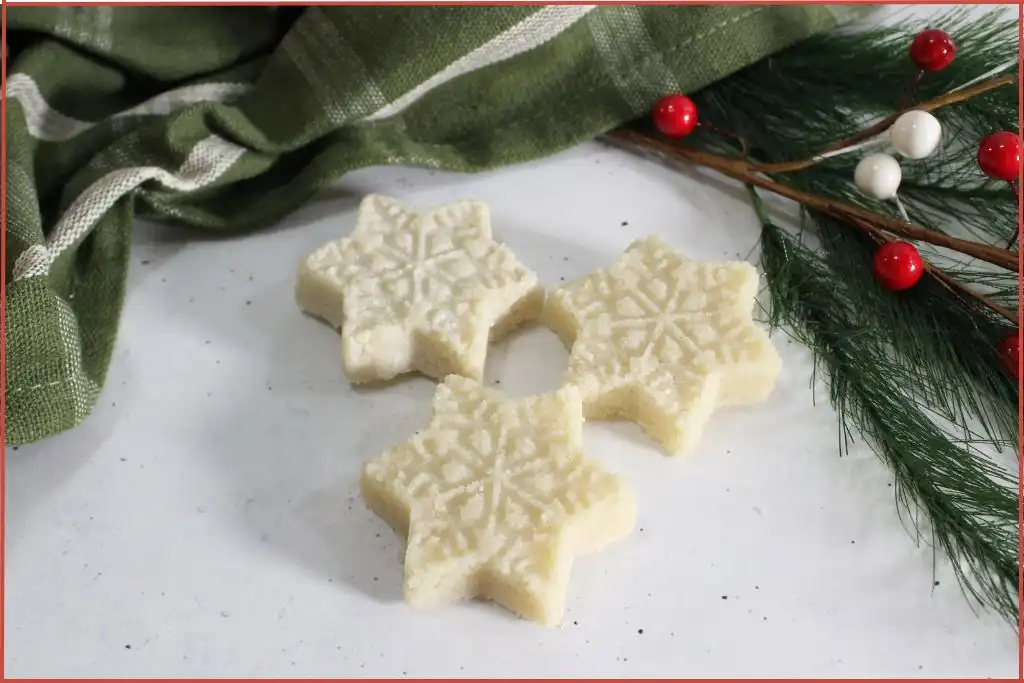 Three white snowflake-shaped sugar scrub bars on a marble surface with evergreen and ribbon, DIY recipe