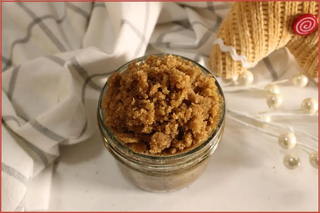 Small glass jar filled with brown gingerbread sugar scrub on a striped kitchen towel, with a knitted gingerbread man decoration wearing a chef hat in the background on a marble tile surface