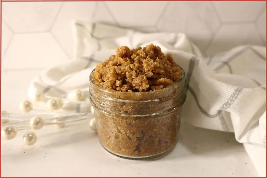 Small glass jar filled with brown gingerbread sugar scrub on a striped kitchen towel on a marble tile surface