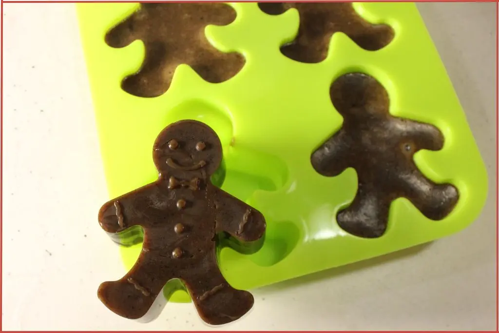 Brown soap shaped like a gingerbread man standing upright in front of a bright green silicone mold filled with more gingerbread man soaps on a light countertop.