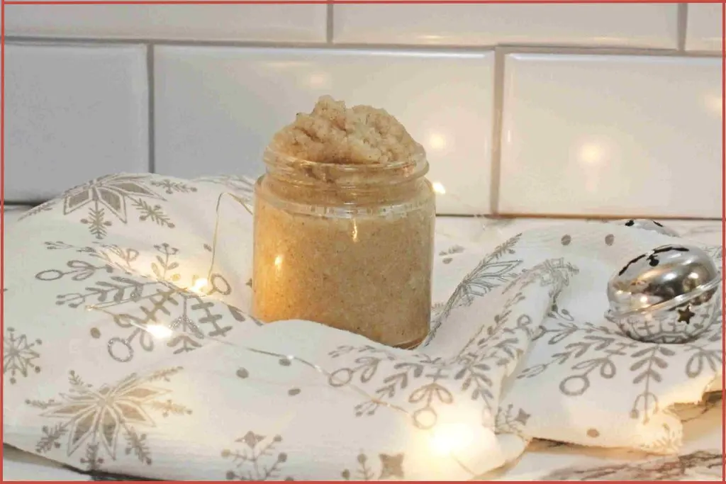 Christmas cookie sugar scrub in a glass jar, sitting on a white and silver snowflake towel with soft holiday lights and a metal ornament in the background