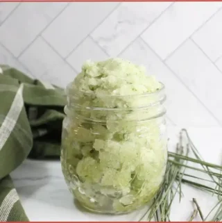 A glass jar filled with a green, homemade Christmas tree sugar scrub with essential oils sits on a white surface; green cloth and pine needles are placed nearby.