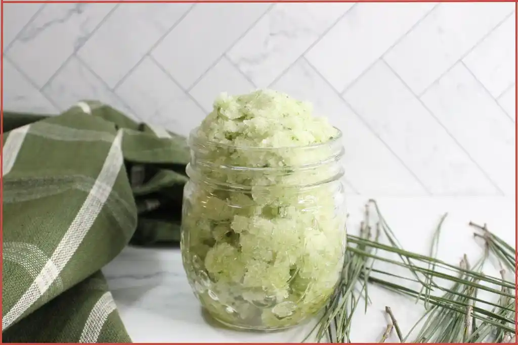 A glass jar filled with a green, homemade Christmas tree sugar scrub with essential oils sits on a white surface; green cloth and pine needles are placed nearby.