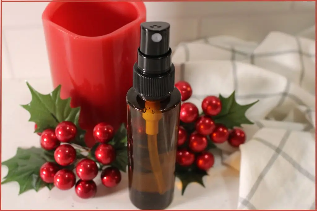 Amber spray bottle of DIY Christmas room spray in front of a red candle, surrounded by faux holly leaves and red berries on a white countertop.