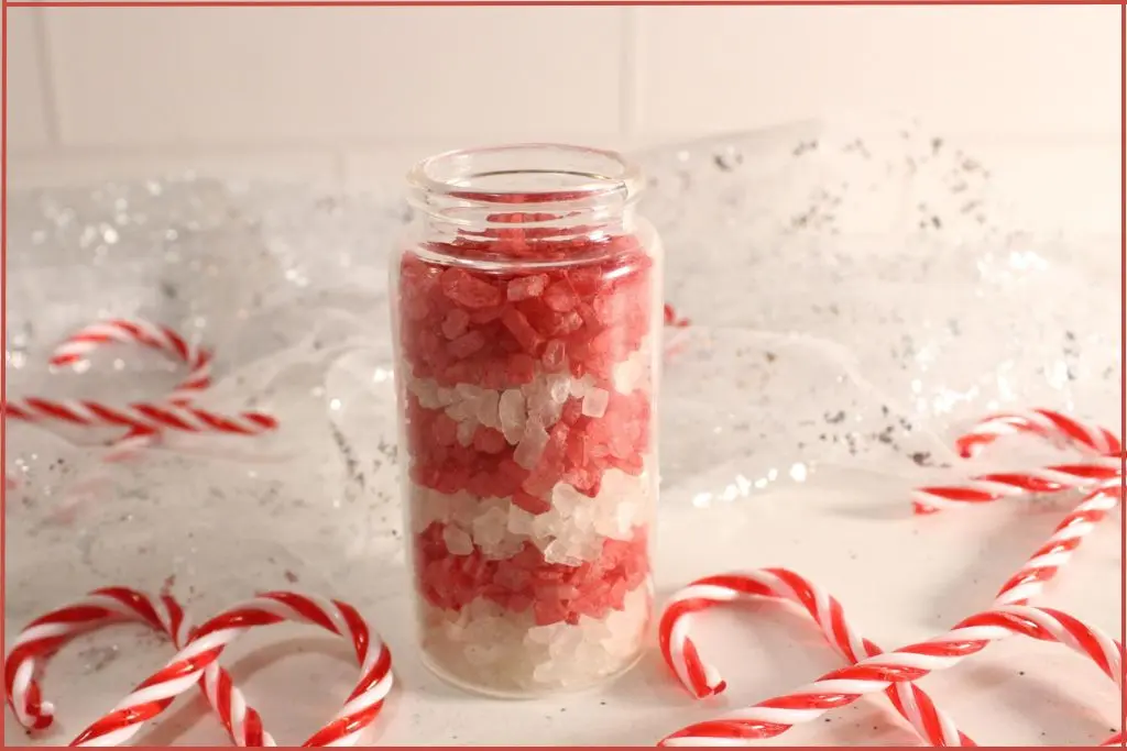 Layered red and white candy cane bath salts in a clear glass jar, surrounded by striped candy canes on a white, sparkly background.