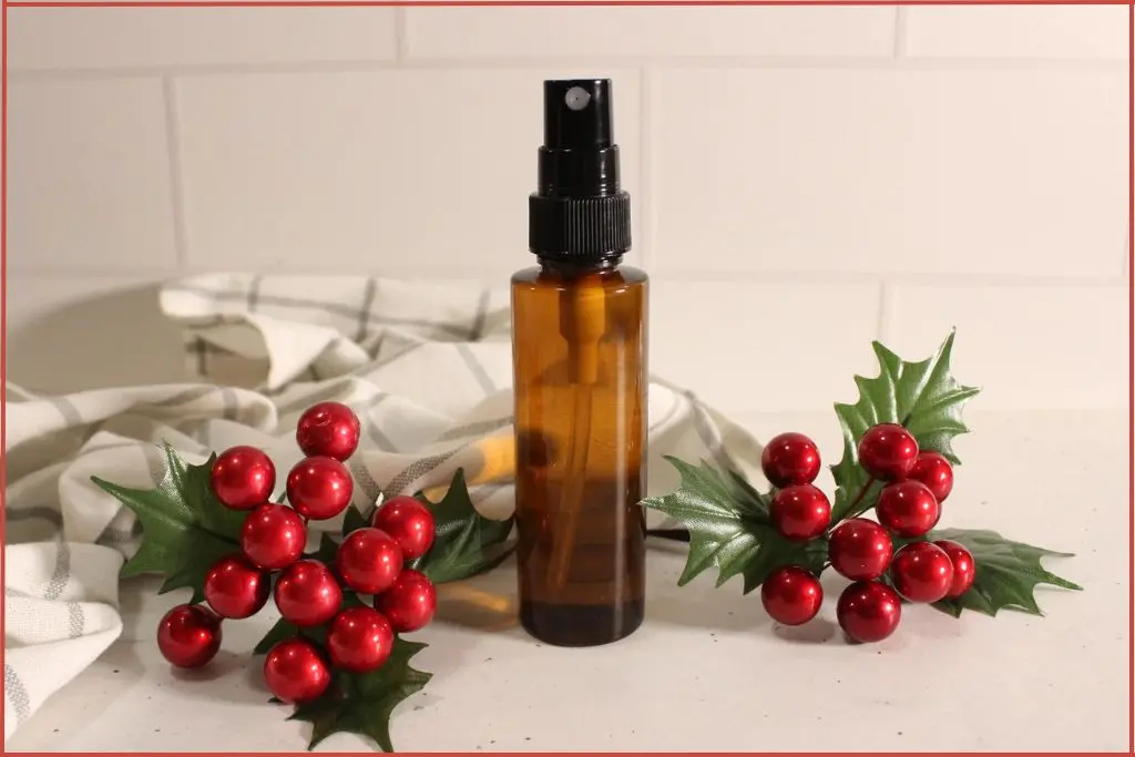 Brown glass spray bottle filled with homemade Christmas room spray, styled with red berry and holly decorations and a white checked towel.