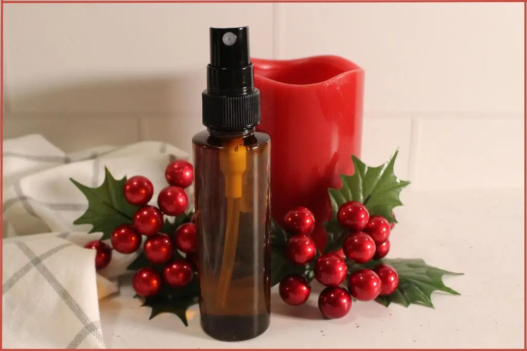 Amber spray bottle of DIY Christmas room spray in front of a red candle, surrounded by faux holly leaves and red berries on a white countertop.
