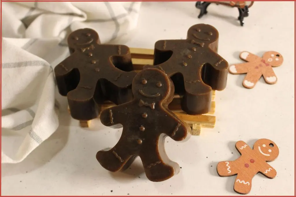 3 brown soaps shaped like gingerbread men on a small wooden rack, styled on a light countertop with a white checkered cloth and two flat gingerbread man decorations.