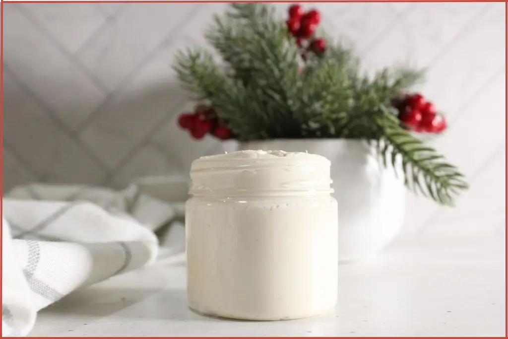 Small glass jar with homemade whipped Christmas hand cream on a light countertop, with a white cloth and a festive evergreen and red berry arrangement in a vase in the background.