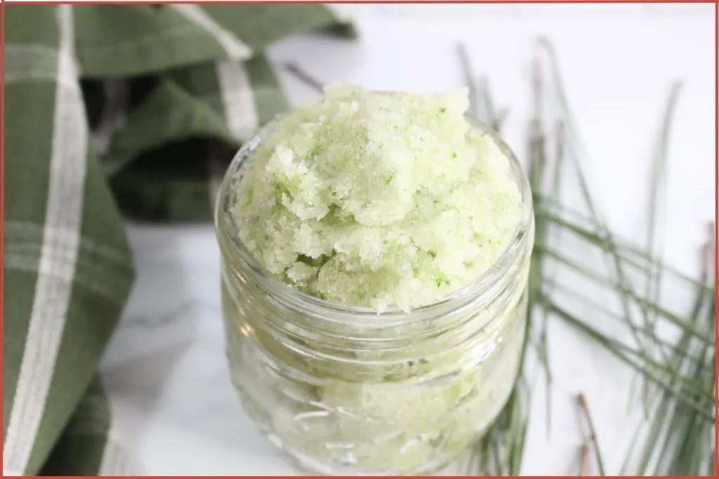 A glass jar filled with a green, homemade Christmas tree sugar scrub sits on a white surface; green cloth and pine needles are placed nearby.