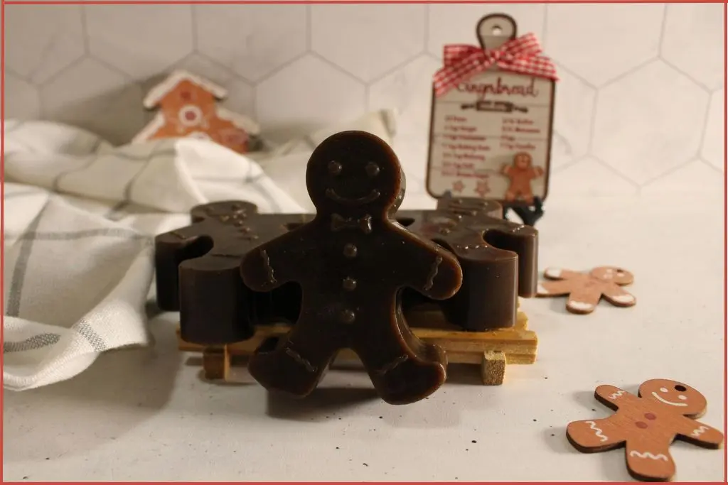 3 brown soaps shaped like gingerbread men on a small wooden rack, styled on a light countertop with a white checkered cloth and two flat gingerbread man decorations.