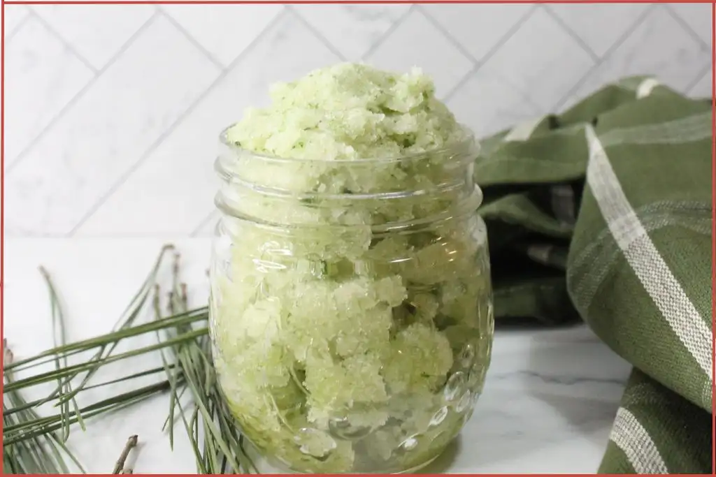 A glass jar filled with a green, homemade Christmas tree sugar scrub with essential oils sits on a white surface; green cloth and pine needles are placed nearby.