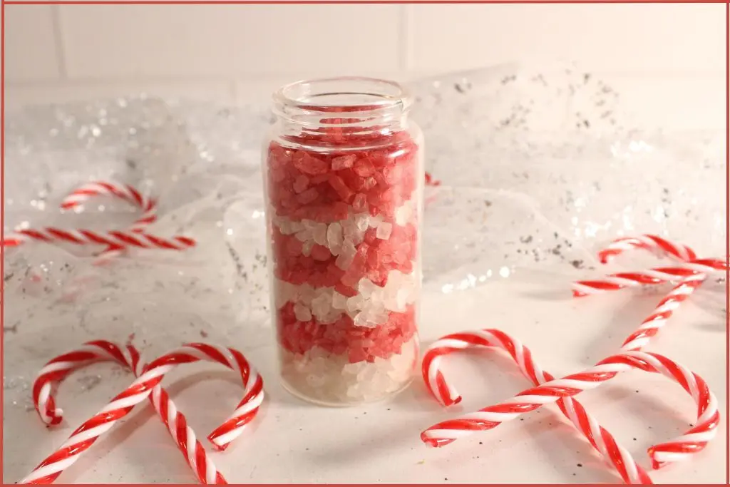 Layered red and white candy cane bath salts in a clear glass jar, surrounded by striped candy canes on a white, sparkly background.