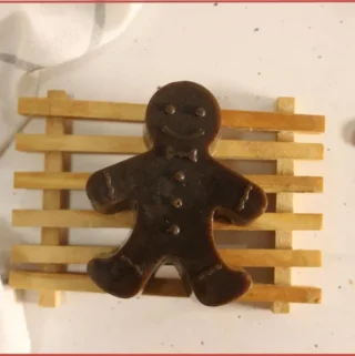 Brown soap shaped like a gingerbread man on a small wooden rack, styled on a light countertop with a white checkered cloth and two flat gingerbread man decorations.