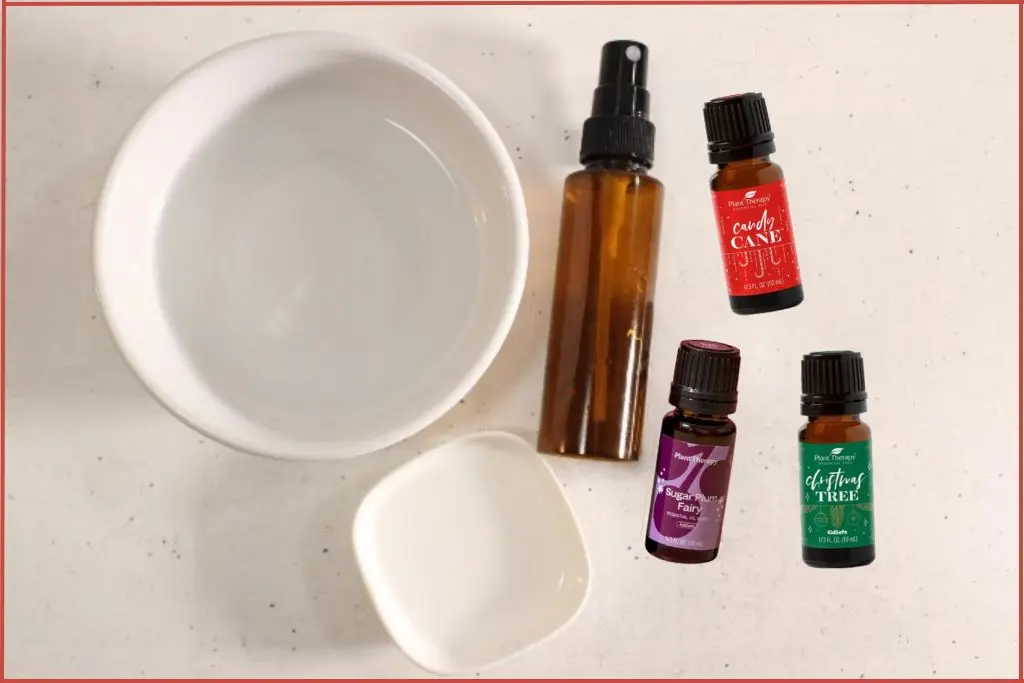 Ingredients to make Christmas room spray - amber spray bottle, bowls with witch hazel and water + 3 bottles of Plant Therapy Christmas essential oil blends