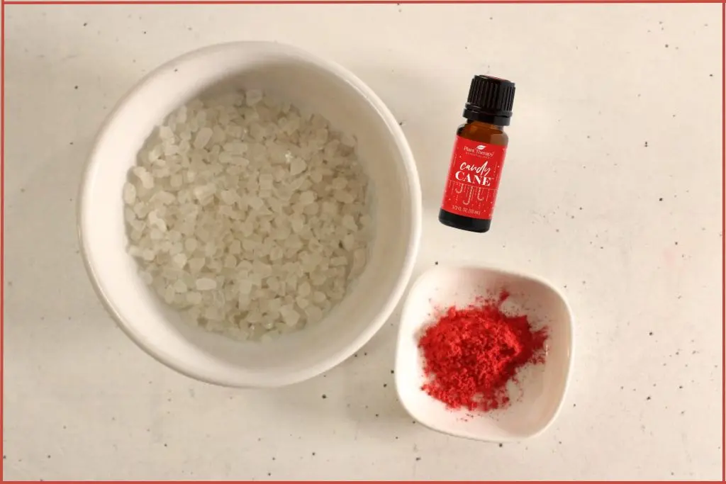 Large bowl of Dead Sea salt, small bowl with red mica powder, and bottle of candy cane blend - ingredients for making candy cane bath salts