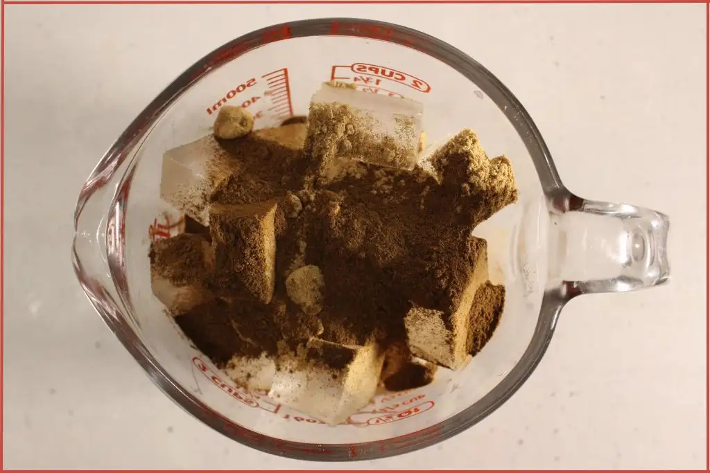 overhead view of glass measuring glass with cinnamon and ginger powder sprinkled on cut melt and pour soap base