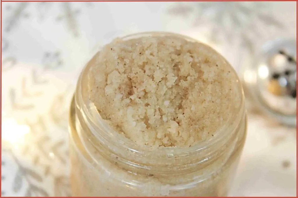 Top view of Christmas cookie sugar scrub in a glass jar, sitting on a white and silver snowflake towel with soft holiday lights and a metal ornament in the background