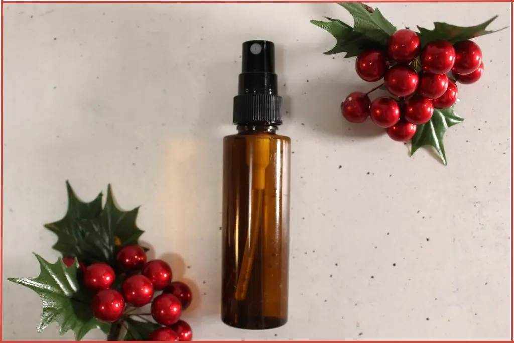 Overhead view of an amber spray bottle of DIY Christmas room spray on a light countertop, framed by red berry and holly leaf decorations.