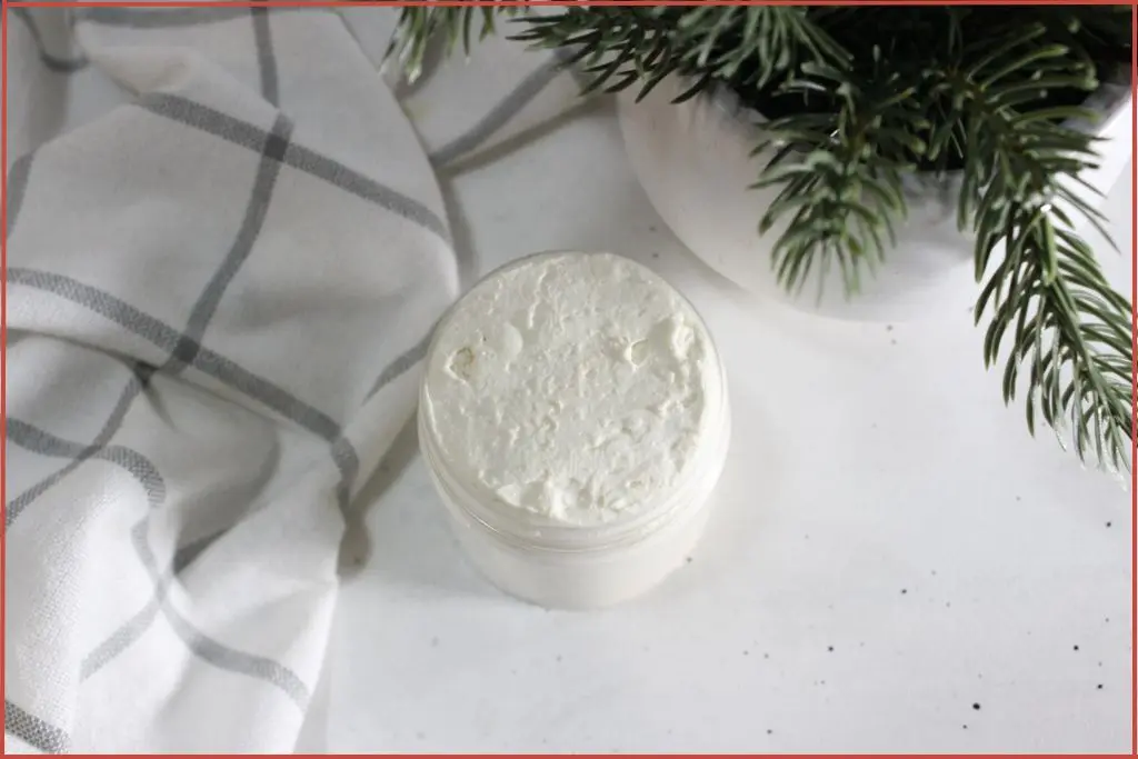 Small glass jar with homemade whipped Christmas hand cream on a light countertop, with a white cloth and a festive evergreen arrangement in a vase in the background.