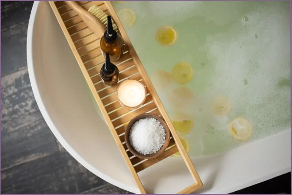 bath tray with two pump bottles and a bowl of bath salts lying across a bath tub filled with water and citrus slices