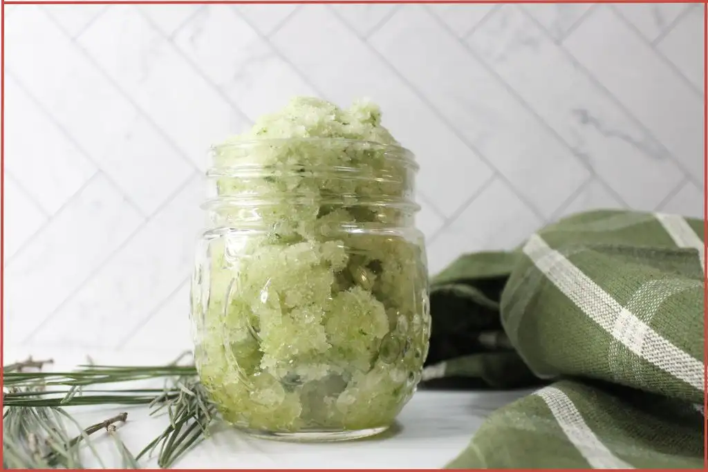 A glass jar filled with a green, homemade Christmas tree sugar scrub sits on a white surface; green cloth and pine needles are placed nearby.