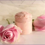 Glass jar of pale pink rose whipped body butter with two pink roses on a white surface